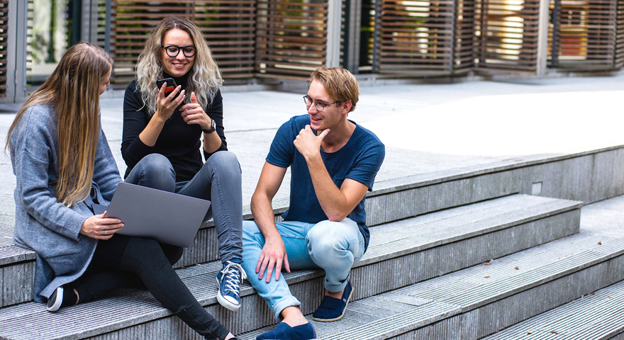 Three persons sitting talking to each other on an outside stair, one holding a laptop