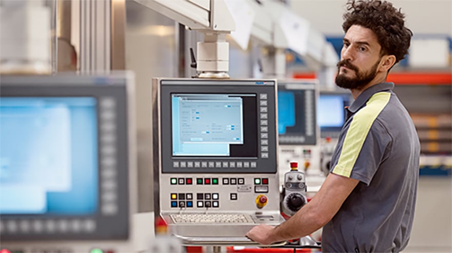 A man in work clothes standing next to a row of monitors and an industrial environment